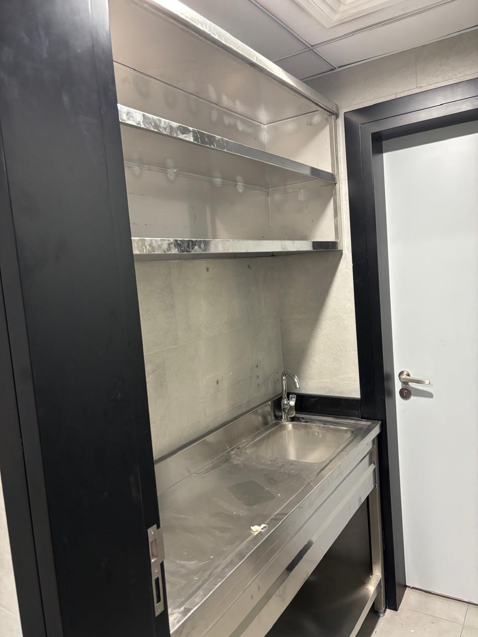 Stainless steel office pantry with counter, sink, faucet, and two open shelves above grey tiled walls